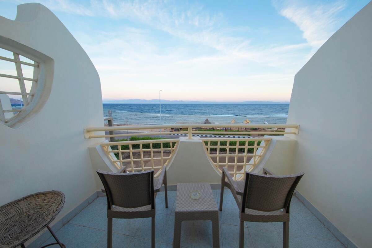 A private balcony is adorned with two wicker chairs and a small table, offering a view of the serene coastline and distant mountains. The beach can be seen below, framed by a soft sky transitioning into twilight.