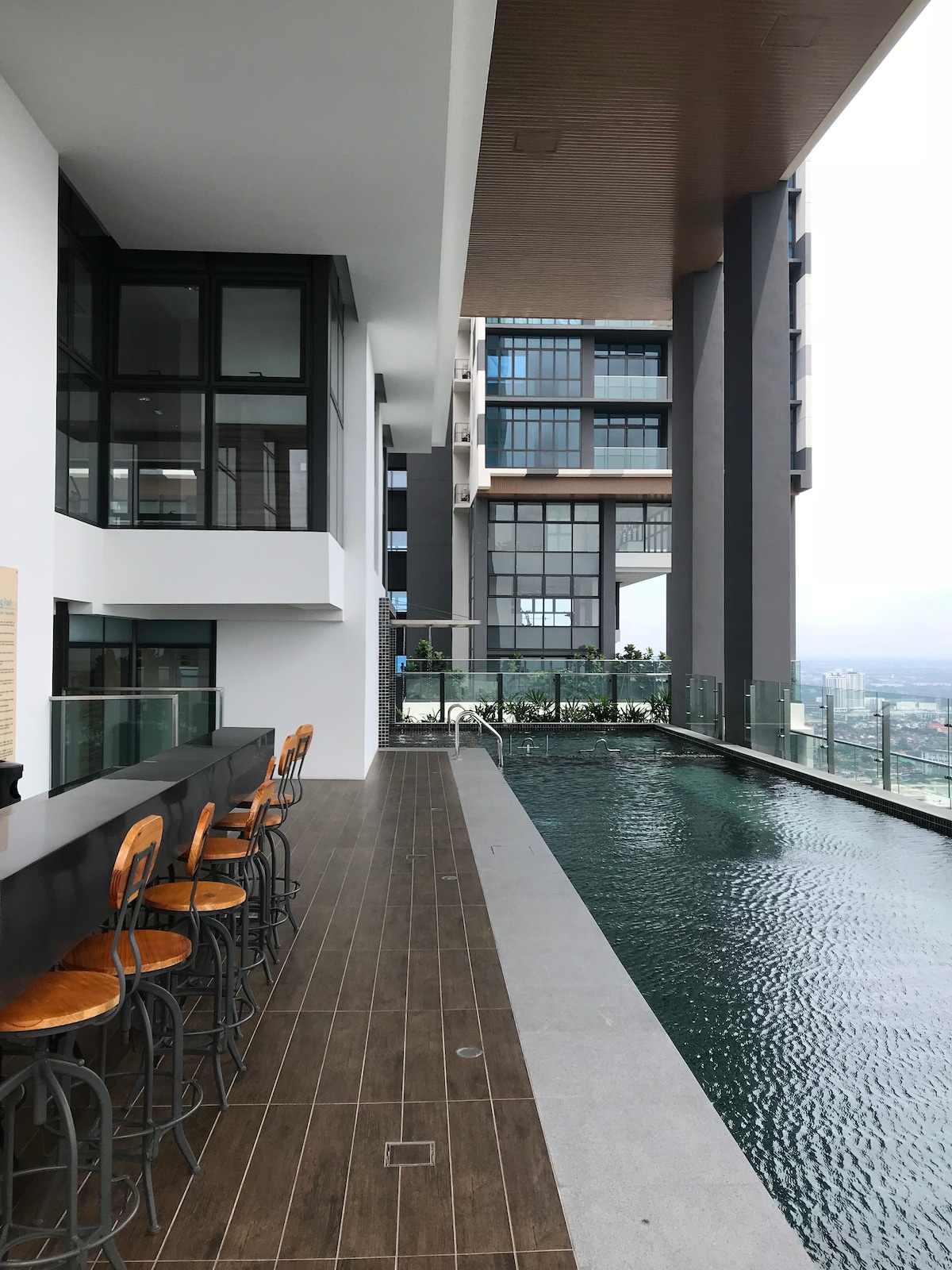 An inviting outdoor space features a sleek bar with high stools alongside a tranquil swimming pool. Glass panels provide an unobstructed view of the surrounding area, while the modern architecture creates a serene and stylish environment.