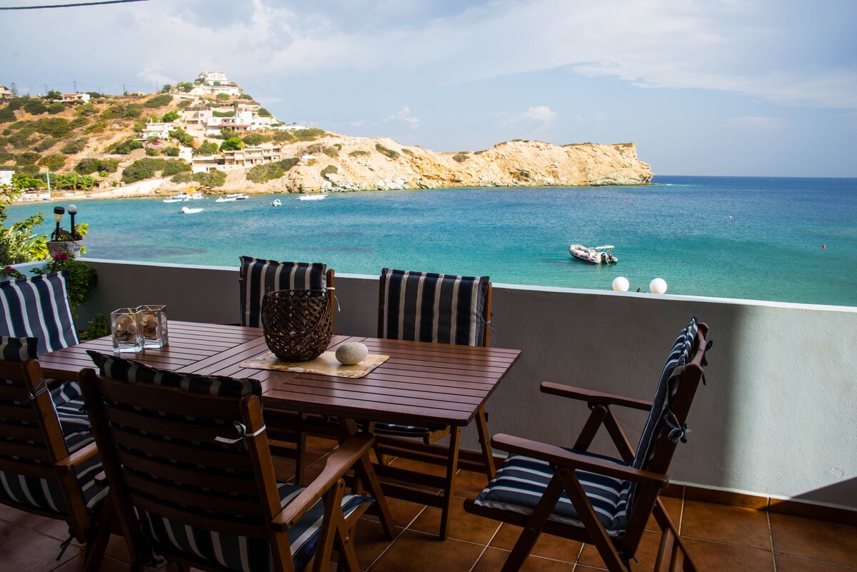 An outdoor dining area is set on a terrace with a wooden table and folding chairs. The sea is visible in the background, with gentle waves lapping at the shore. A rocky coastline and distant hills are framed by clouds in the sky, creating a serene seaside view.