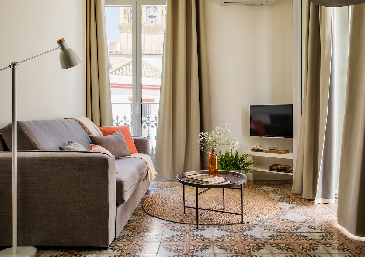 A cozy living area features a gray sofa adorned with colorful pillows. A round coffee table sits on a patterned tiled floor, accompanied by a decorative vase. Large windows are framed by flowing curtains, allowing natural light to illuminate the space.