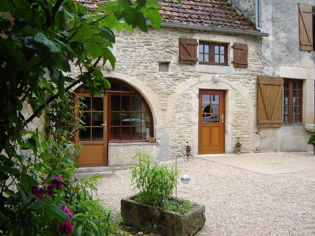 The exterior of a rustic farmhouse features a stone façade with wooden shutters and double doors. Lush greenery and colorful flowers enhance the entrance, while a gravel path leads to the house. An arched window adds character to the inviting atmosphere.