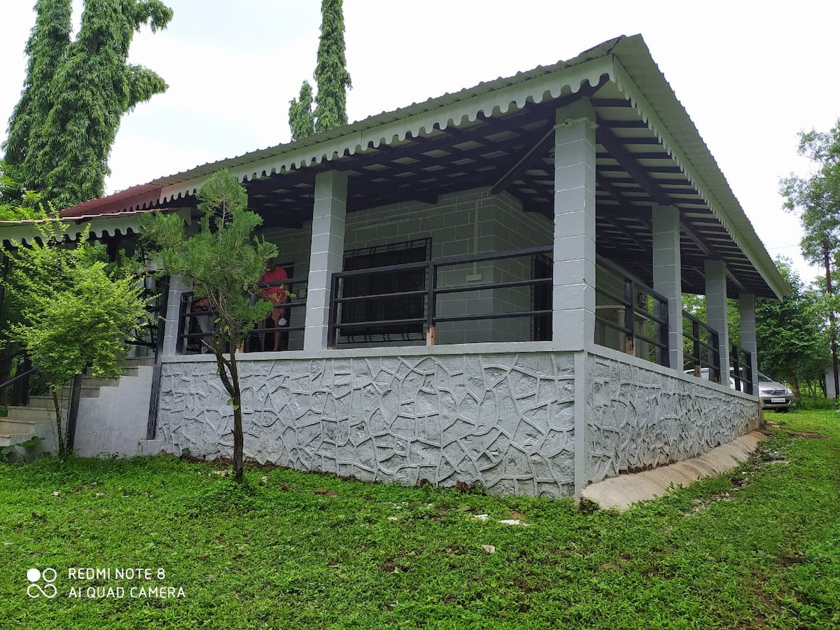 The exterior of the house is showcased, featuring a stone wall and a spacious balcony. Lush green grass surrounds the home, with tall trees providing shade. The structure has a sloping roof and large windows, offering a welcoming view of the tranquil setting.