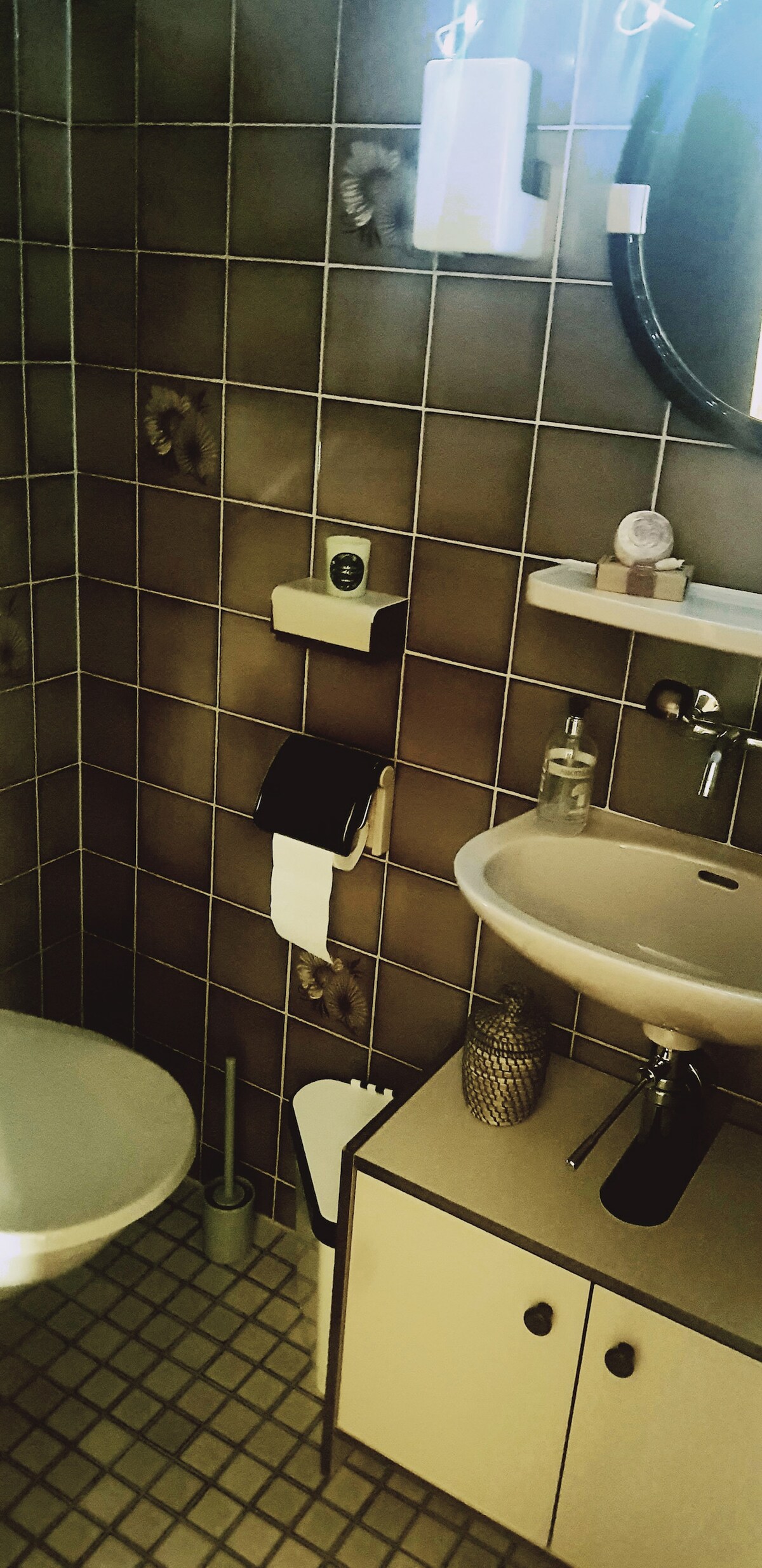 A bathroom with tiled walls features a round mirror above a modern sink. An assortment of toiletries is organized neatly on a shelf. A toilet is positioned beside a waste bin, and a decorative element adds a touch of style to the space.