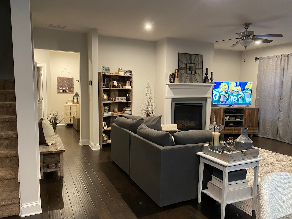 A comfortable living area features a gray sectional sofa positioned facing a wall-mounted television. A modern fireplace serves as a focal point. Warm hardwood floors contrast with white walls, while a bookcase filled with various items completes the inviting space.
