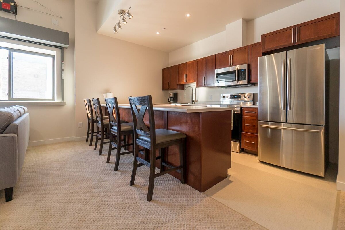 A modern kitchen area features dark wood cabinetry and stainless steel appliances, including a refrigerator and oven. A spacious island with seating for six is present, alongside a window allowing natural light to brighten the room. An inviting living area complements the kitchen.