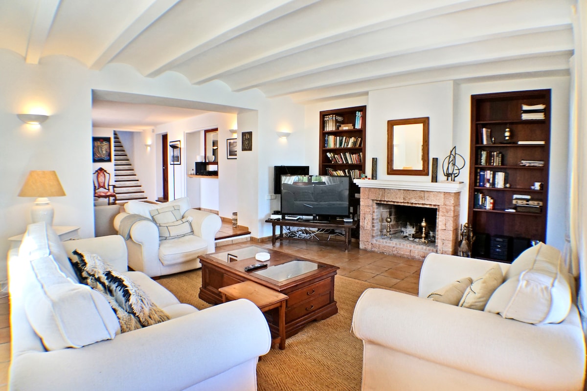 A spacious living room features cream-colored sofas arranged around a wooden coffee table. A flat-screen television is situated on a console, while a fireplace complements the room's ambiance. Bookshelves line the walls, filled with various books, and soft lighting adds to the inviting feel.