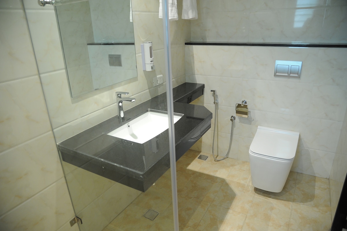 A modern bathroom features sleek marble tiles with a spacious walk-in shower. A white sink is paired with a dark countertop, and a large mirror reflects the minimalistic design. The contemporary toilet is situated beside the sink, creating a streamlined appearance.