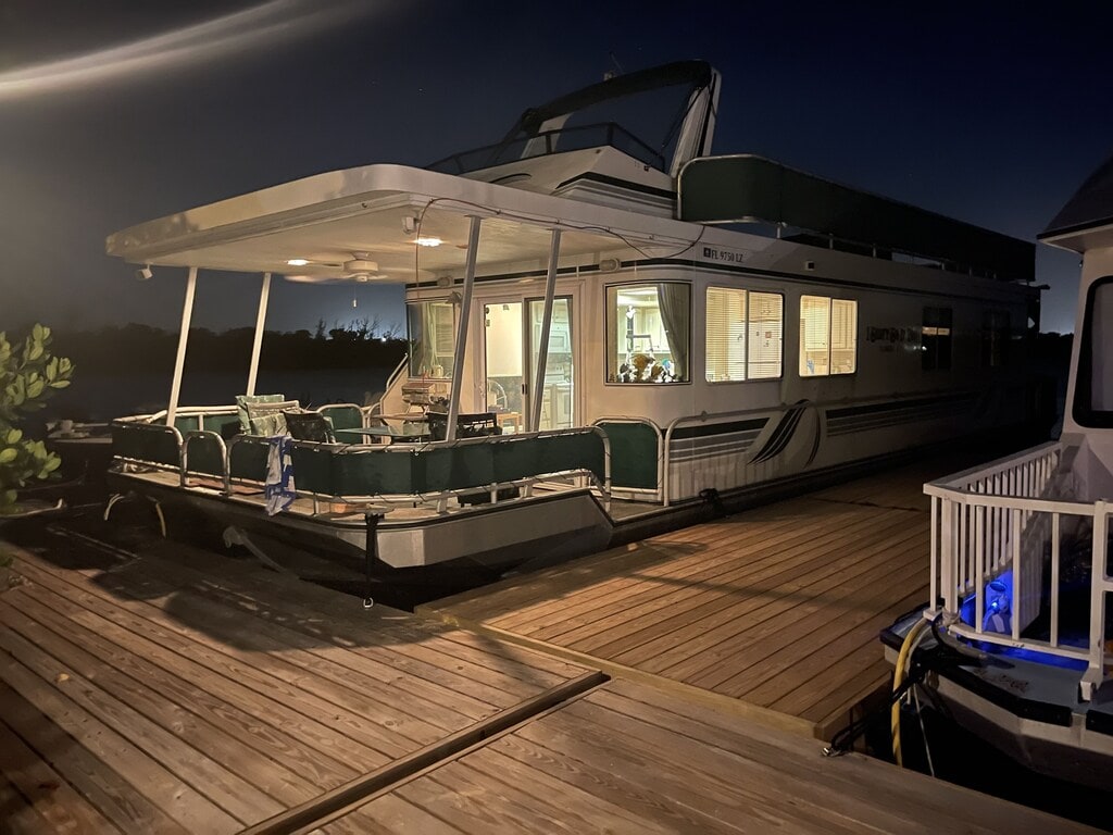 The houseboat is illuminated at night, displaying a spacious covered patio with multiple chairs. Glass doors provide glimpses into the living space. The structure is docked on a wooden platform, surrounded by the calm water, creating a serene evening setting.