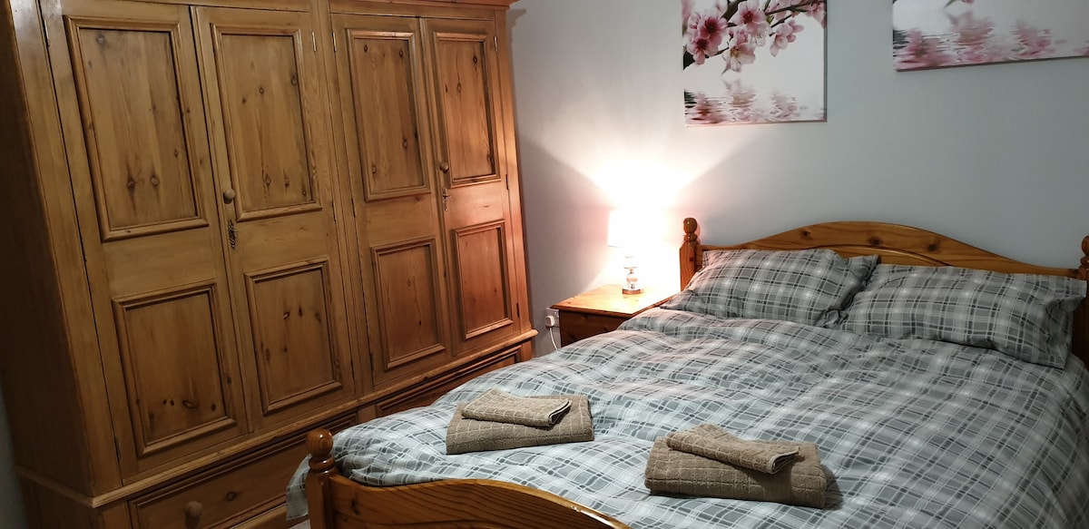 A cozy guest room features a spacious double bed with a plaid duvet, complemented by two neatly arranged towels. An oak wardrobe provides ample storage, while a bedside table holds a lamp, offering soft lighting. Floral artwork adorns the wall, enhancing the room's decor.