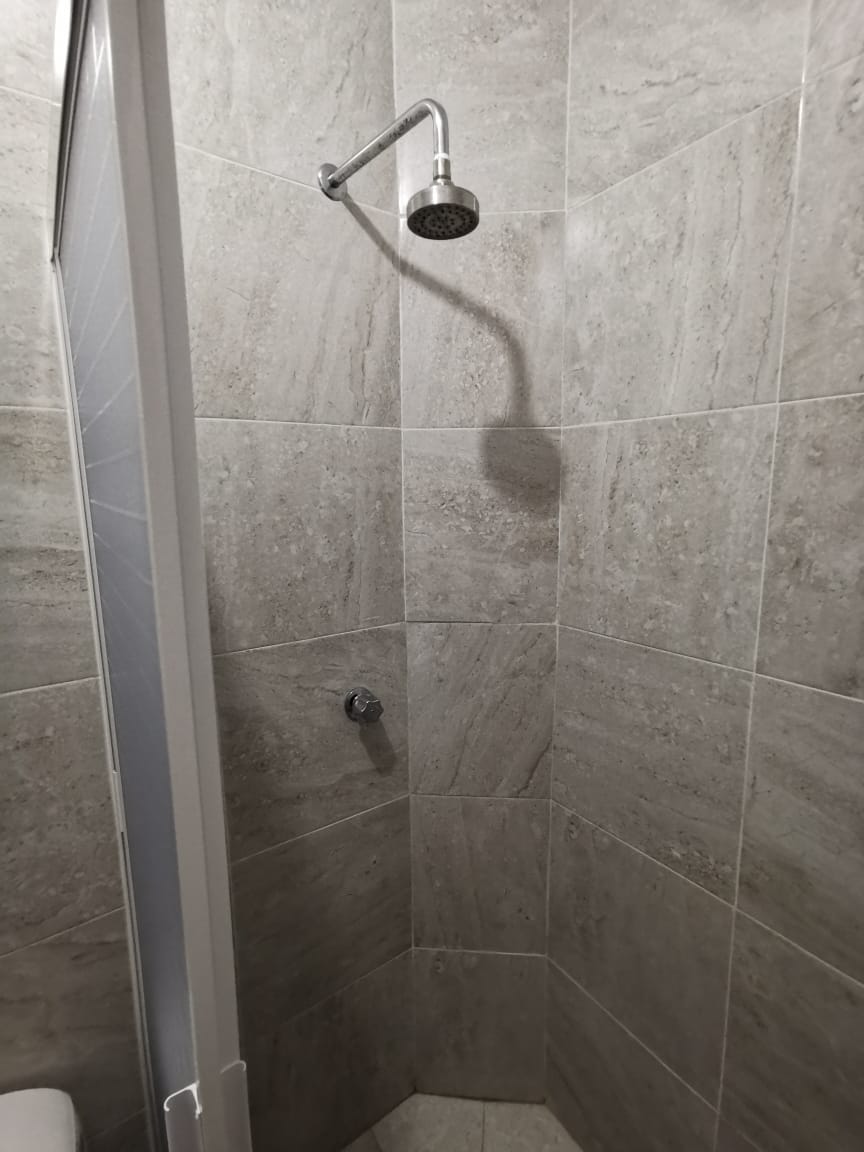 A corner shower space is enclosed by clear glass. The shower features a modern showerhead mounted on a tiled wall with neutral grey tones. The seamless design offers a clean and contemporary aesthetic.