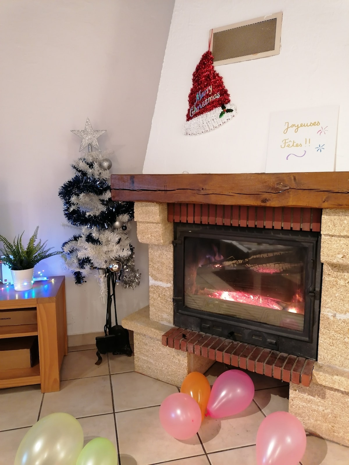 A cozy fireplace is set within a stone surround, creating a warm ambiance. A small, decorated Christmas tree and festive decorations are placed nearby. Colorful balloons are scattered on the tiled floor, adding a cheerful touch to the space.