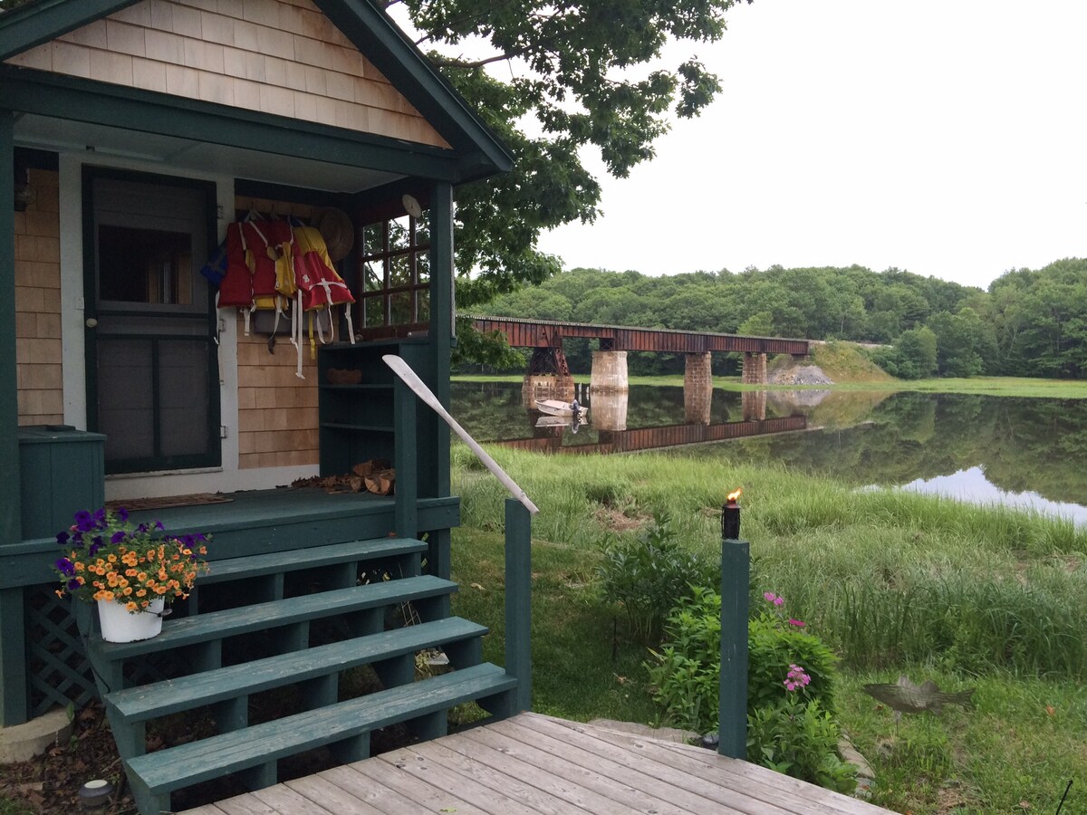 A welcoming porch features a flower pot filled with vibrant blooms and life jackets hanging nearby. The view from the porch looks over a calm river reflecting greenery, with a rustic bridge visible in the distance, enhancing the serene riverside setting.