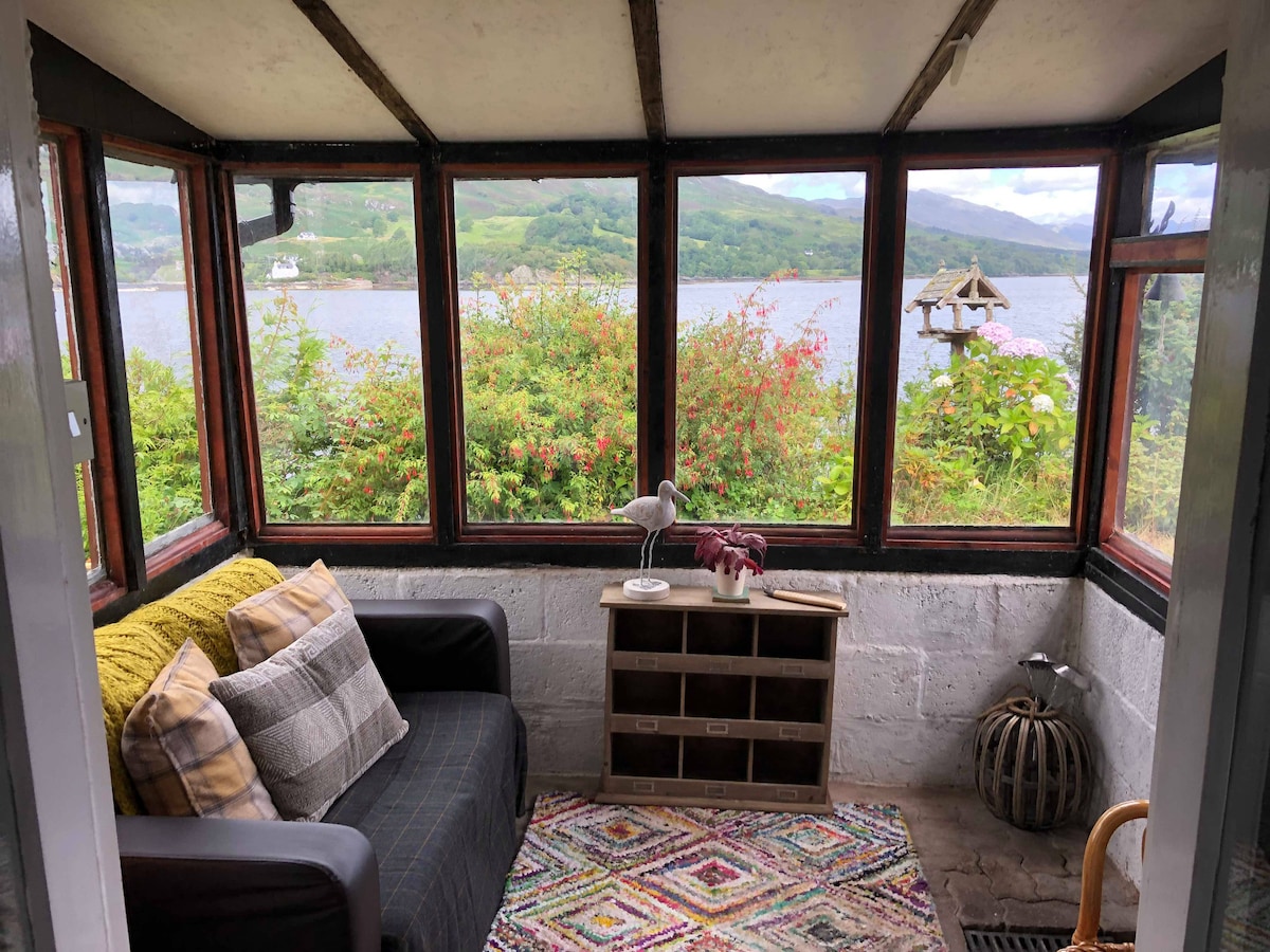 A sunlit sitting area features large windows providing views of the loch and surrounding greenery. A cozy couch with decorative cushions is paired with a small lamp and a wooden storage unit. A colorful rug enhances the inviting atmosphere.