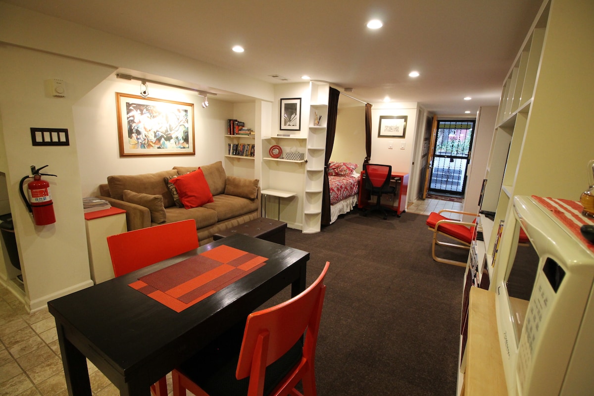 An inviting studio space is seen with a comfortable sofa adorned with red cushions, a dining table with red chairs, and a cozy sleeping area with fresh bedding. Soft overhead lighting illuminates the warm interior, creating a welcoming ambiance.