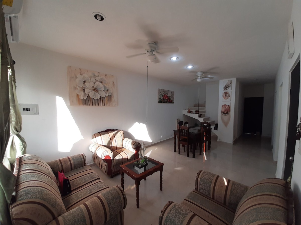 A well-lit living area features a pair of patterned sofas and a wooden coffee table, complemented by a small green plant. A dining table is visible in the background, surrounded by chairs, while artwork adorns the white walls.