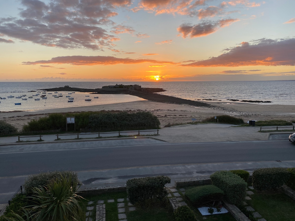 A serene sunset over the beach is captured, featuring a vibrant sky with hues of orange and pink. The calm waters reflect the colors, while small boats are visible on the horizon. The foreground shows a pathway lined with greenery, leading towards the shore.
