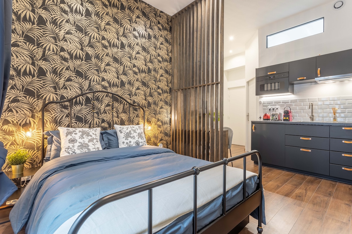 A spacious sleeping area features a double bed with a blue coverlet and decorative pillows. Behind the bed, a patterned wallpaper adds texture, while a wooden divider hints at separate living space. Bright light filters in from a window, illuminating the modern kitchen area in the background.