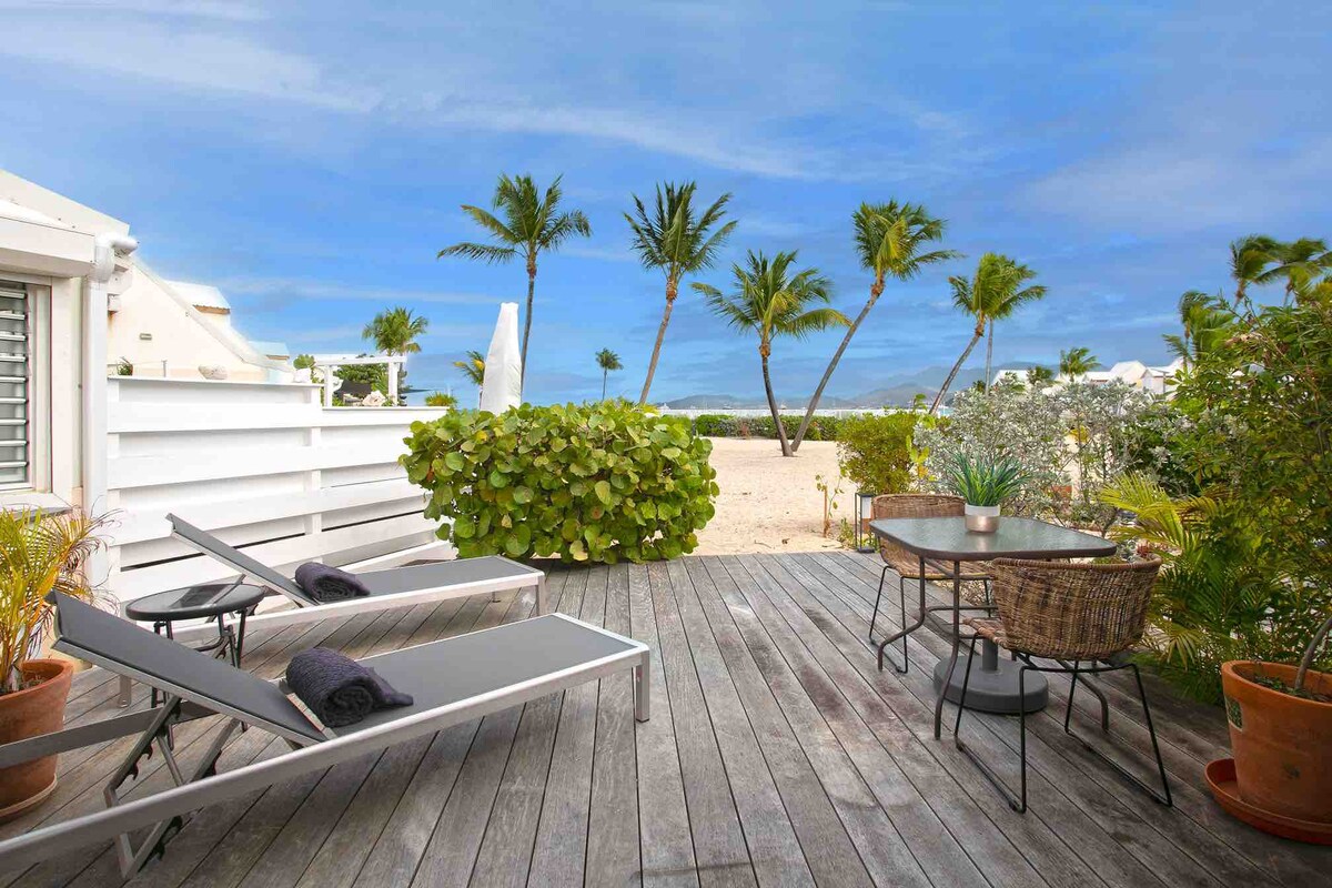 A wooden terrace features two lounge chairs positioned for relaxation, accompanied by a round dining table and woven chairs. Lush greenery and palm trees surround the area, with soft sand visible beyond, creating a serene outdoor space. The sky displays a calm shade of blue.