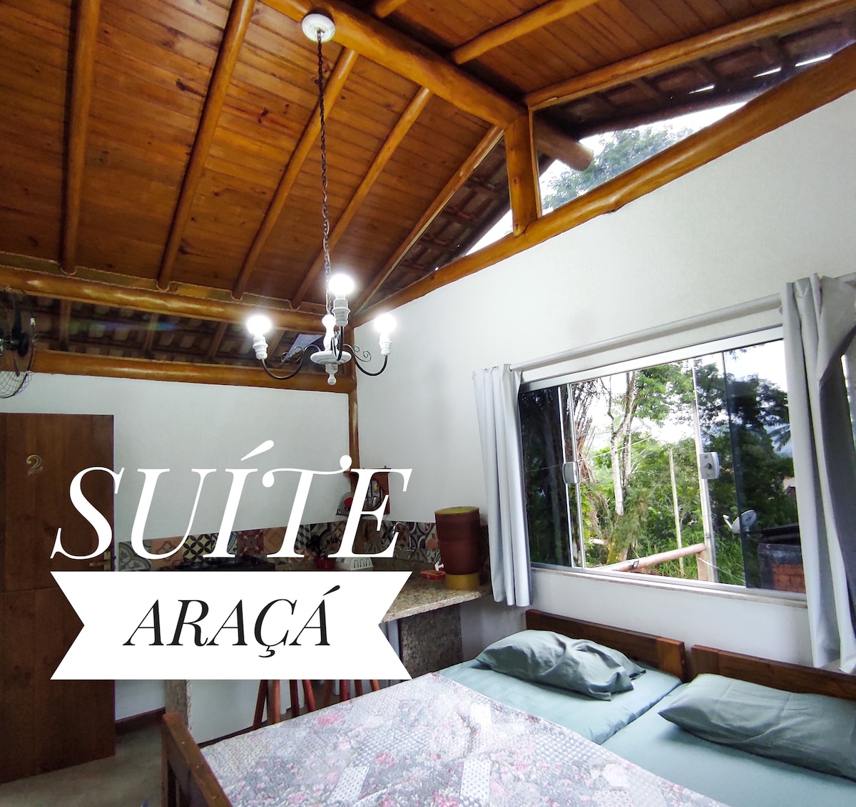 The suite features a spacious sleeping area with a wooden ceiling and large windows allowing natural light to fill the room. A table is set nearby, complemented by comfortable bedding on the bed. Greenery is visible outside the windows, enhancing the tranquil atmosphere.