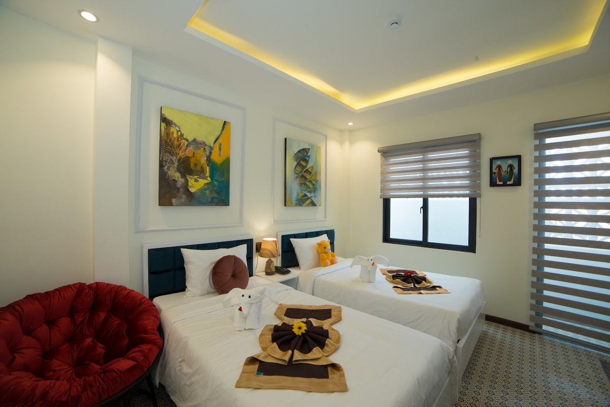 A well-lit guest room features two twin beds, each adorned with decorative towels. Colorful artwork hangs on the walls, while a circular lounge chair adds texture to the space. A large window with blinds provides natural light, complementing the modern design.