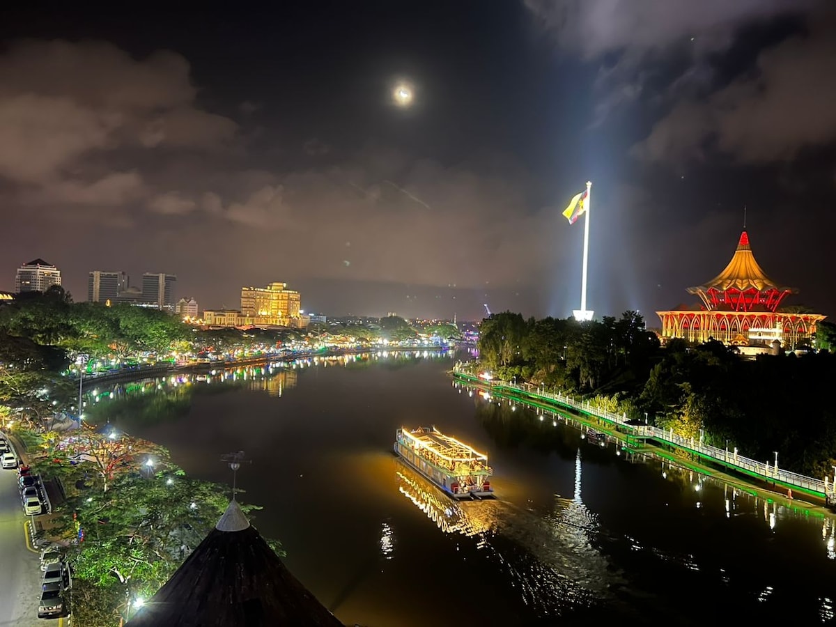 The night view captures a serene river scene illuminated by lights. A boat is seen gently gliding across the water, while the backdrop features prominent buildings and the majestic State Legislative Assembly, enhancing the vibrant atmosphere along the waterfront.