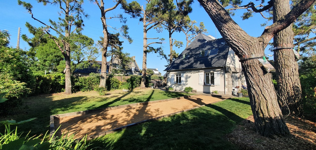 A spacious garden is surrounded by tall trees, providing a serene environment. A designated pétanque court can be seen in the foreground, featuring sand and bordered by greenery. The house, featuring a sloped roof and multiple windows, stands peacefully in the background.