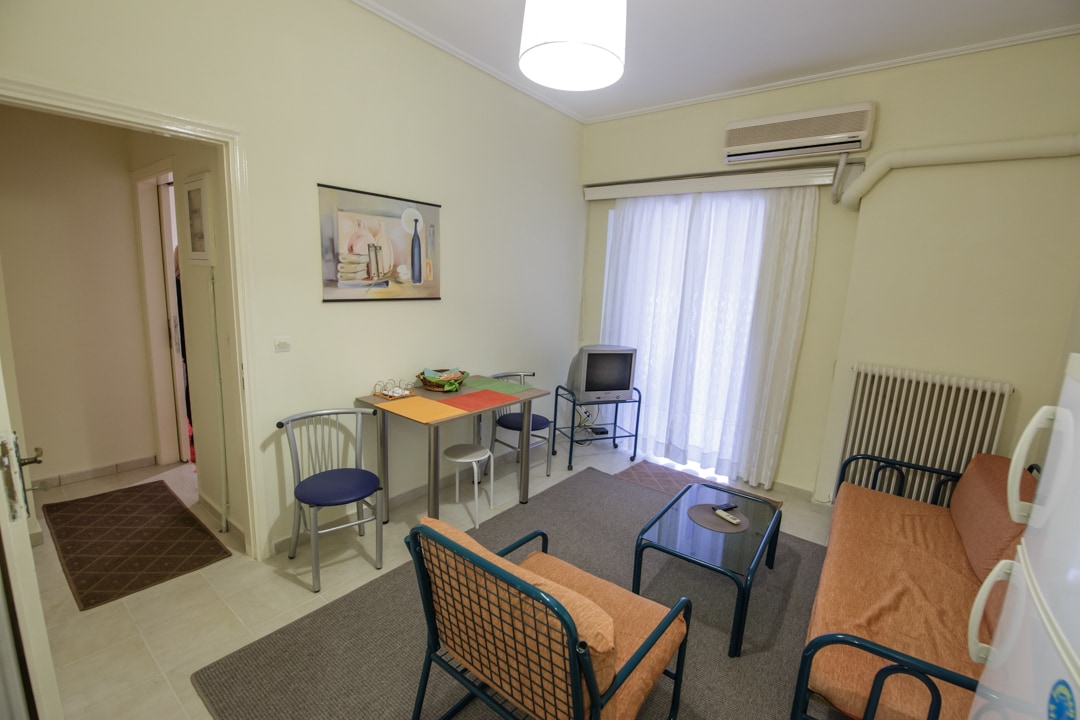 A cozy living area features a beige rug on tiled flooring. Two comfortable sofas and a pair of armchairs surround a small dining table. A television stands in the corner, with light filtering through sheer curtains. The space is air-conditioned with a wall-mounted unit.