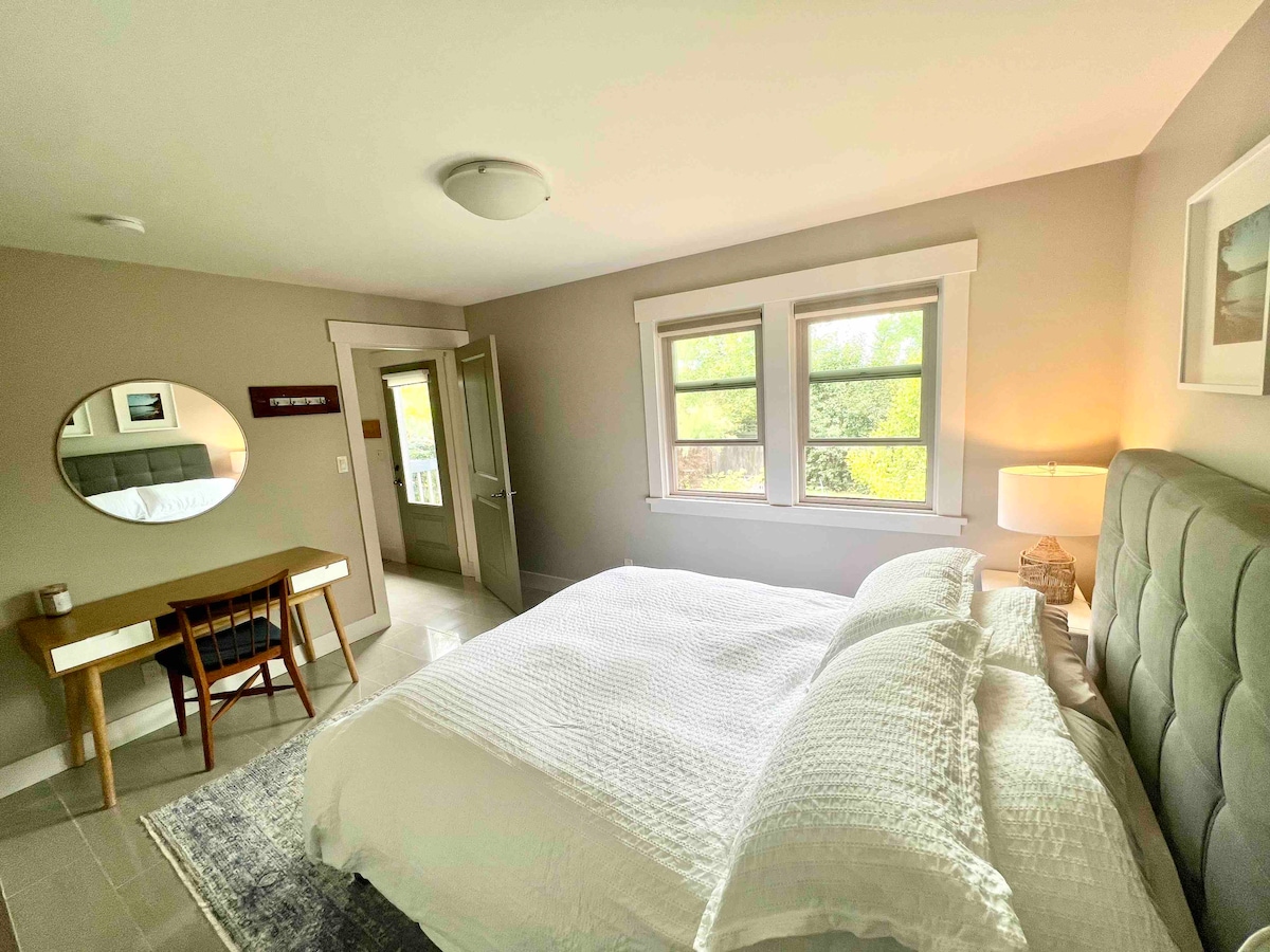 A modern bedroom features a spacious bed with white bedding against a soft gray wall. A round mirror above a desk reflects natural light from a window, while a bedside lamp adds a warm touch. A door leads to an outdoor space, enhancing accessibility.