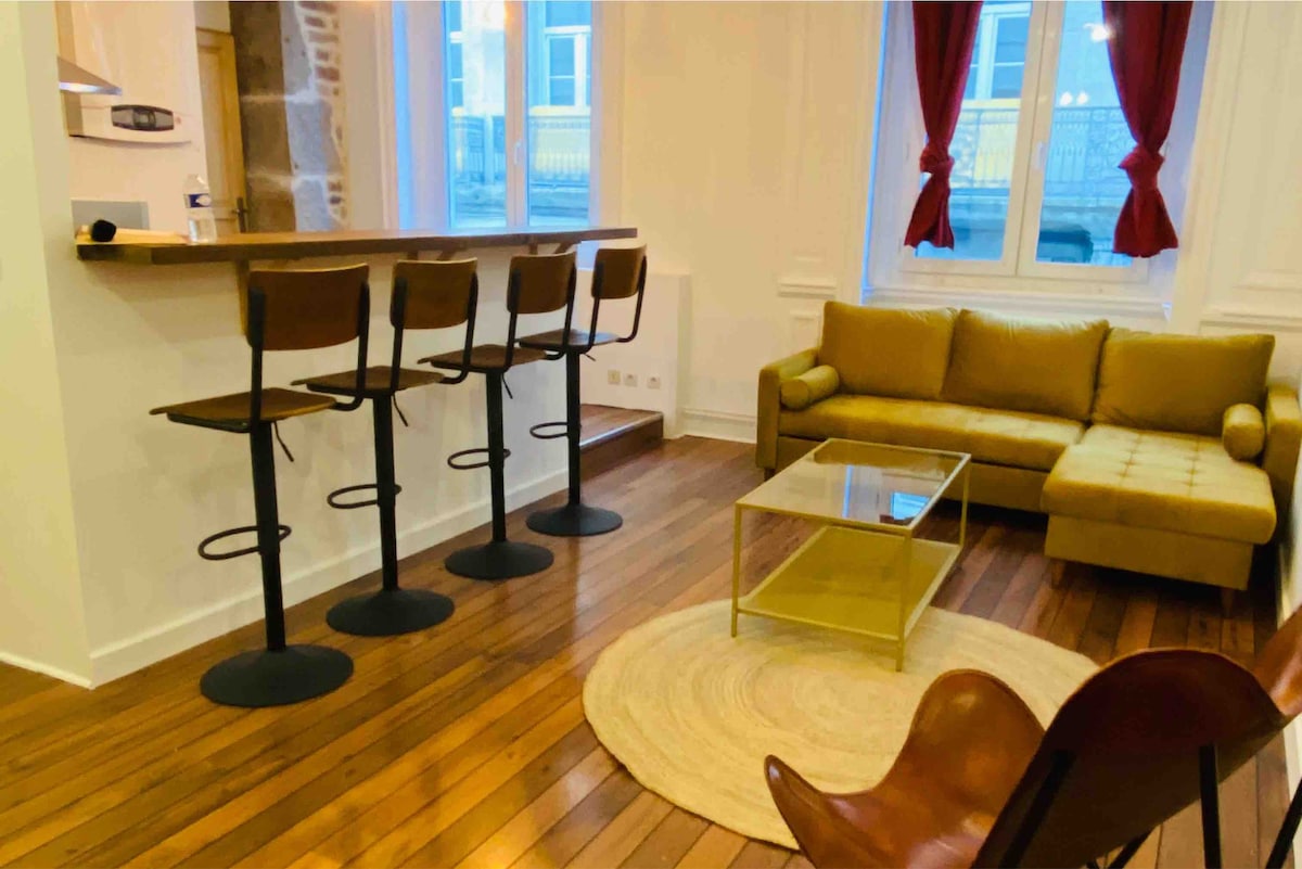 A compact living area features a comfortable sectional sofa in a soft beige color, paired with a glass coffee table. Four high-backed bar stools are positioned along a counter, while natural light streams through large windows with red curtains, highlighting warm wooden flooring.