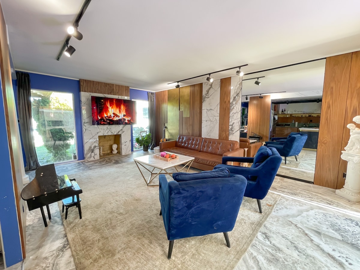 A spacious living area features a mix of modern and classic decor, with a leather sofa and vibrant blue armchairs arranged around a coffee table. A television is mounted above a decorative fireplace, and large windows allow natural light to illuminate the marble flooring.