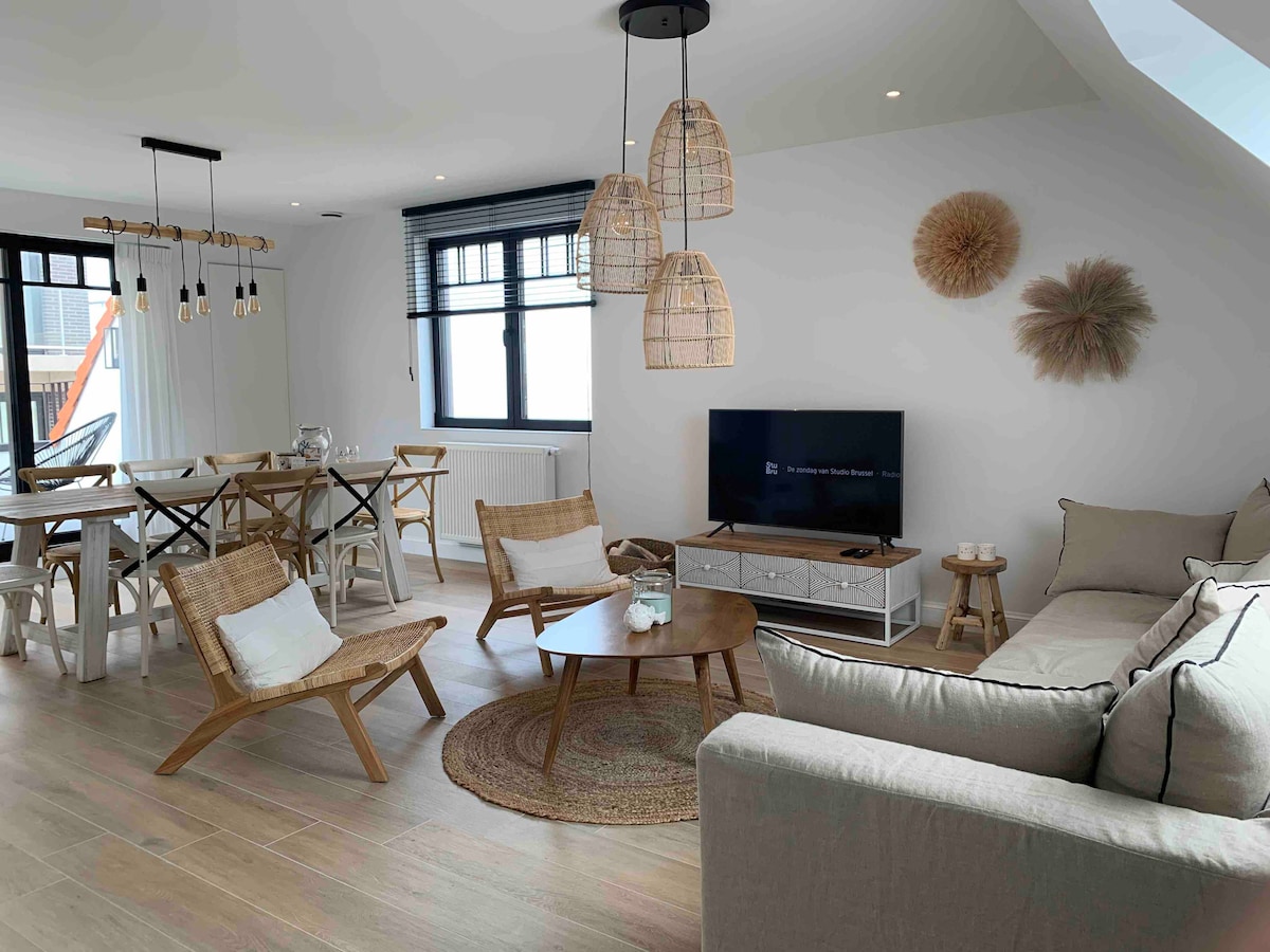 A bright living area features a comfortable sofa and two woven chairs arranged around a circular coffee table. A television is placed on a low cabinet, while a dining table with four chairs is visible in the background. Natural light enters through large windows, enhancing the space.