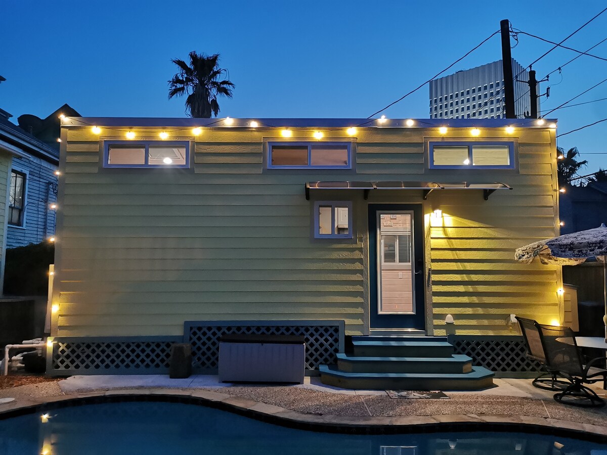 The outdoor view of a tiny retreat is illuminated by soft string lights, highlighting the vibrant yellow exterior. Clear glass windows offer glimpses into the interior. Nearby, a pool reflects the evening sky, creating a serene ambiance in the outdoor space.