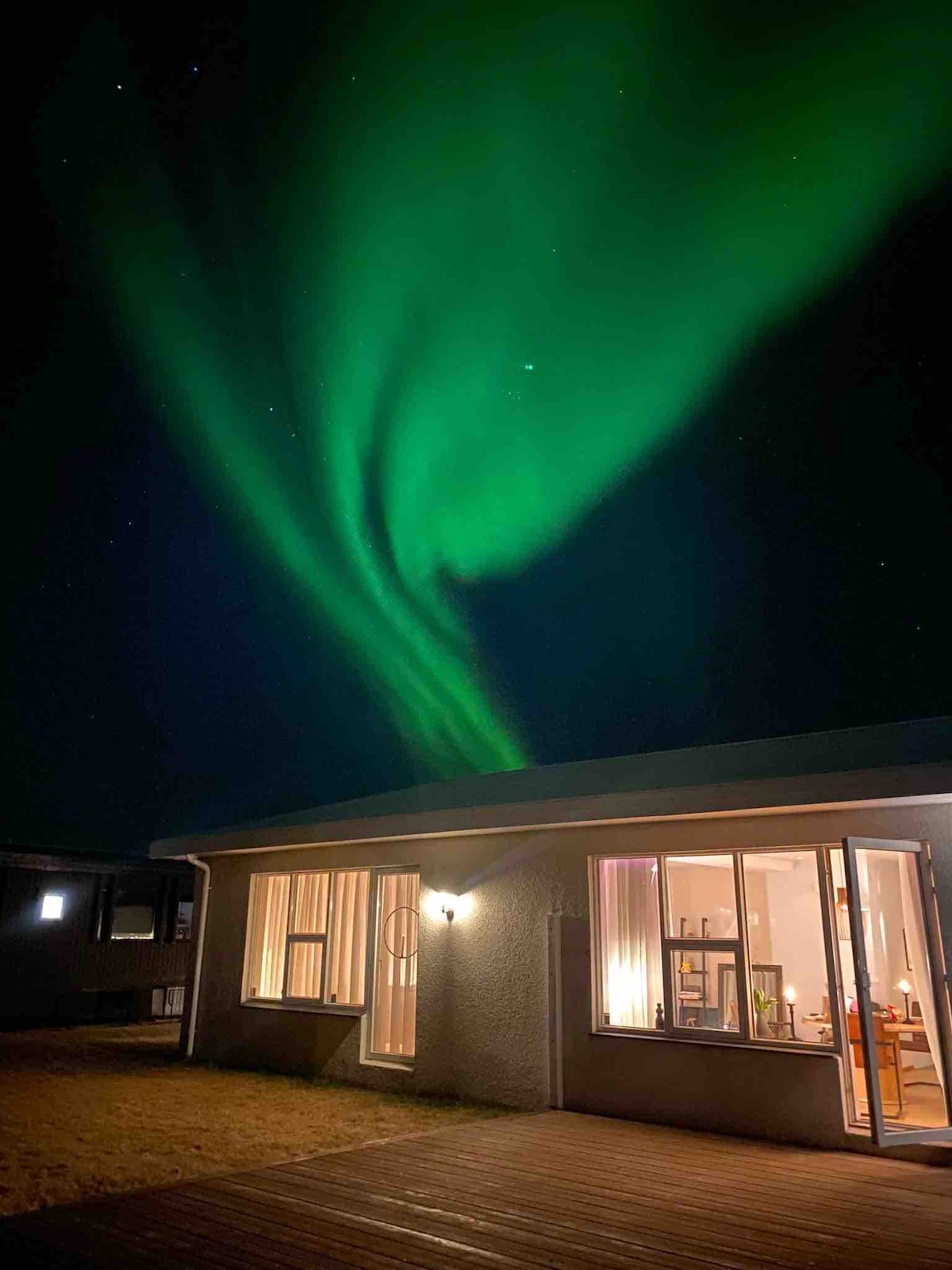A warm glow emanates from the windows of a modest apartment, framing a vibrant display of the Northern Lights swirling in the dark sky above. The outdoor area features wooden decking, enhancing the connection to nature.