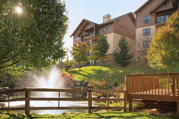 A tranquil outdoor scene is depicted, featuring a gently flowing fountain in a pond surrounded by lush greenery. The three-story building stands in the background, framed by trees displaying autumn foliage, adding a sense of serenity to the environment.