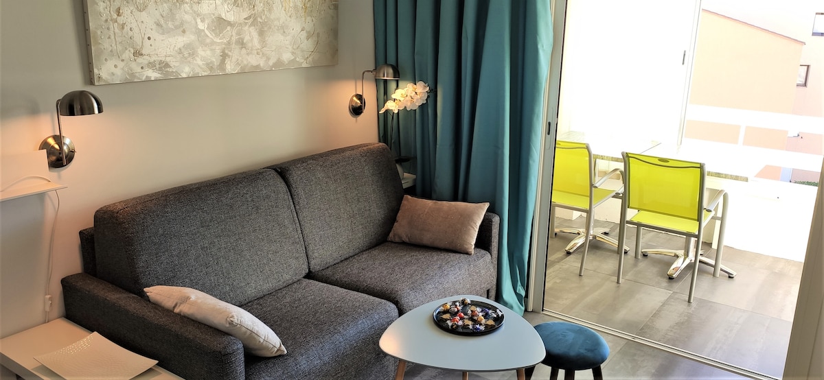 A modern living area features a gray sofa accompanied by a small blue stool and a circular coffee table. Two wall-mounted lamps provide lighting. The room opens to a loggia with outdoor seating visible through sliding glass doors, introducing a sense of spaciousness and light.