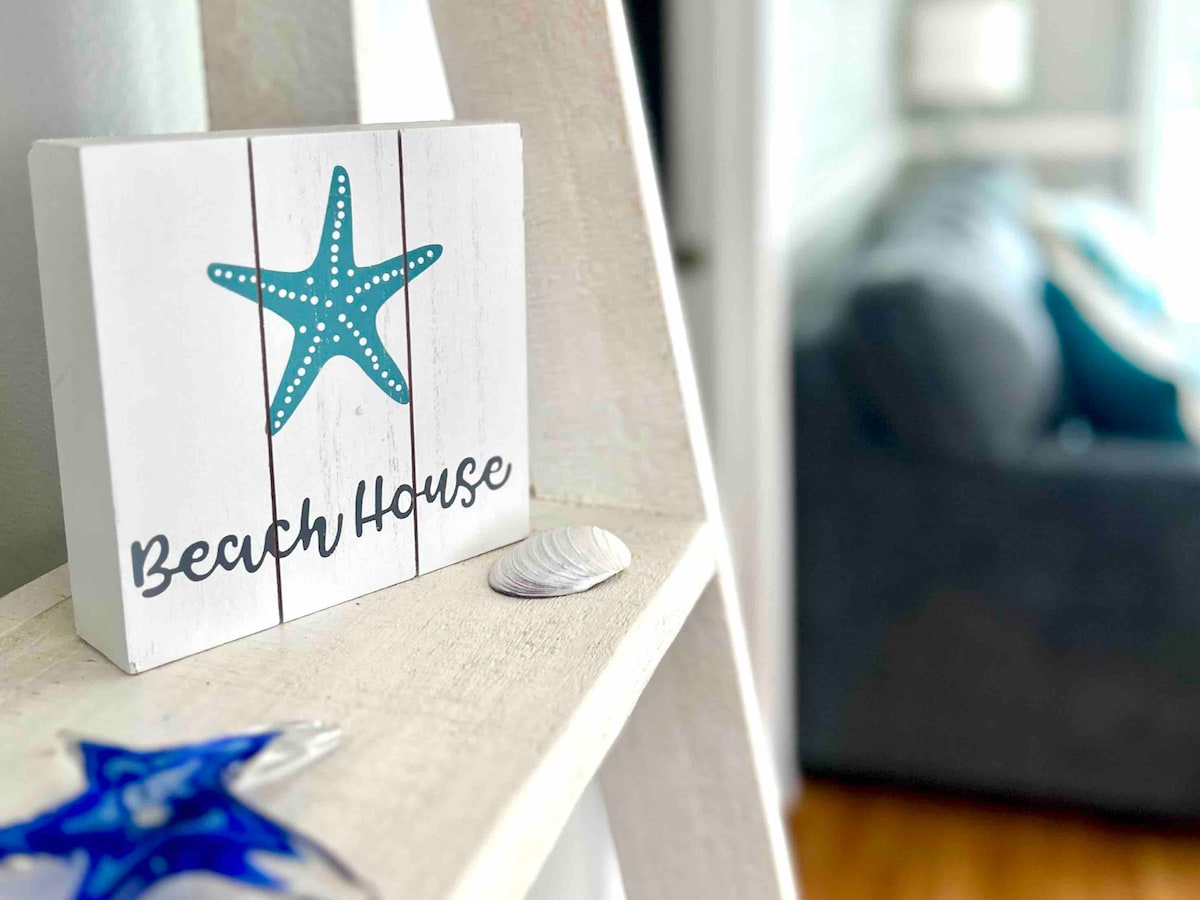 A decorative sign featuring a teal starfish and the words 'Beach House' is displayed prominently on a shelf. A light-colored shell sits beside it. The background shows a cozy living area with a gray sofa and light blue cushions.