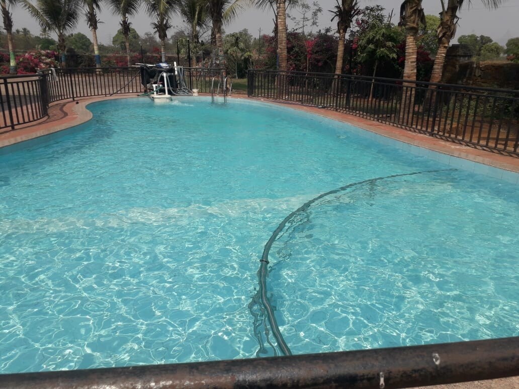A large swimming pool is surrounded by a wrought iron fence, featuring clear blue water. Palm trees and colorful flowering plants can be seen in the background, contributing to a serene outdoor setting.