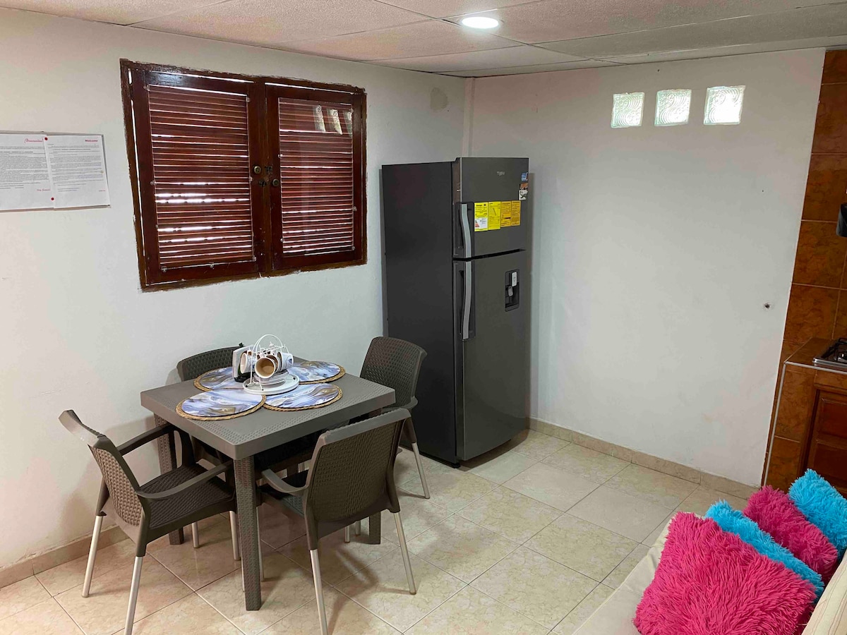 A functional kitchen area is highlighted by a dining table set for four, with plates and utensils neatly arranged. A modern refrigerator stands against the wall, while plush cushions in vibrant colors add a touch of comfort to the adjacent seating area.