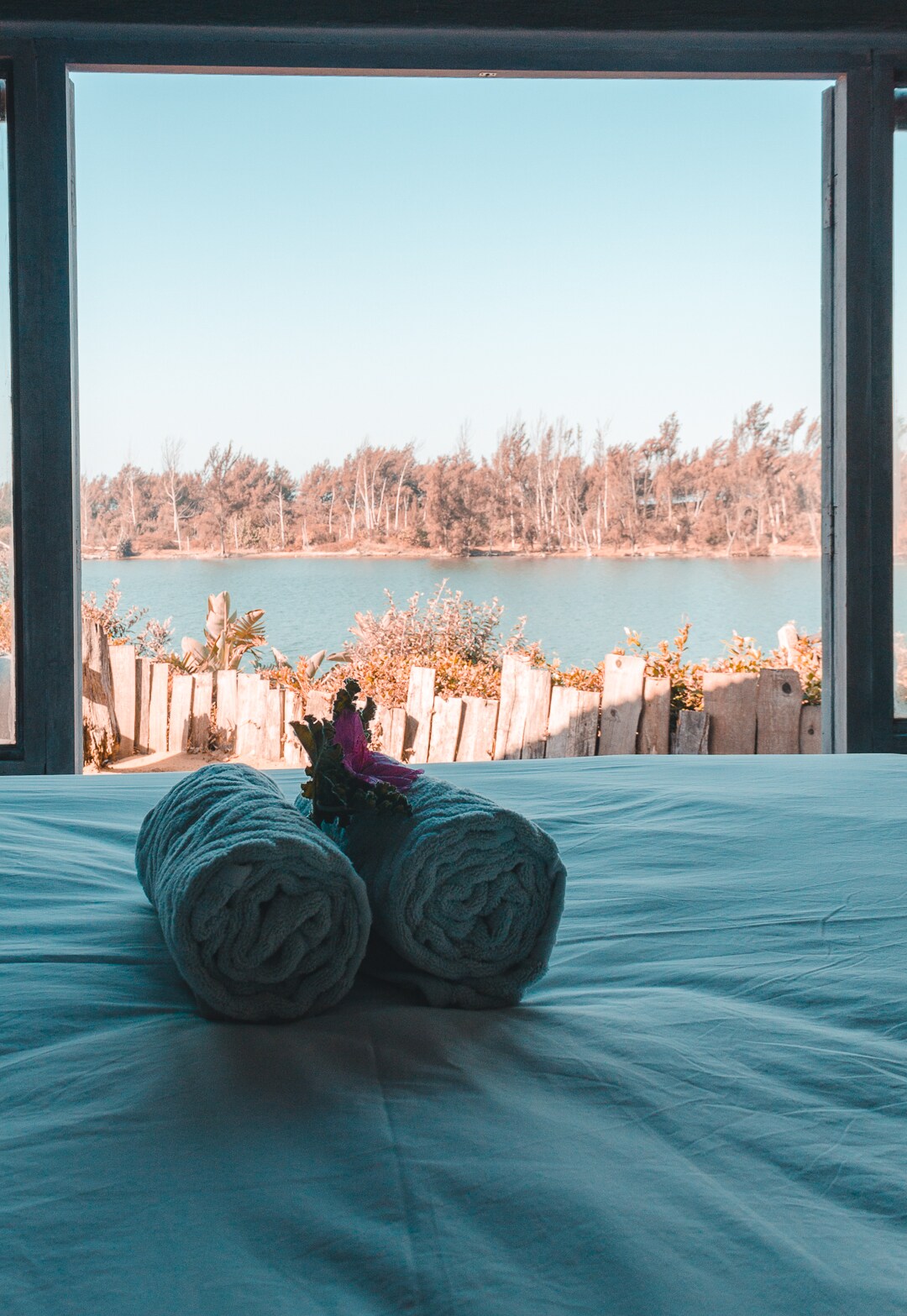 A spacious bed is positioned near a large window showcasing views of the lagoon and surrounding greenery. Rolled towels are neatly arranged at the forefront, adding a touch of comfort. Natural light fills the room, enhancing the connection to the serene outdoor landscape.