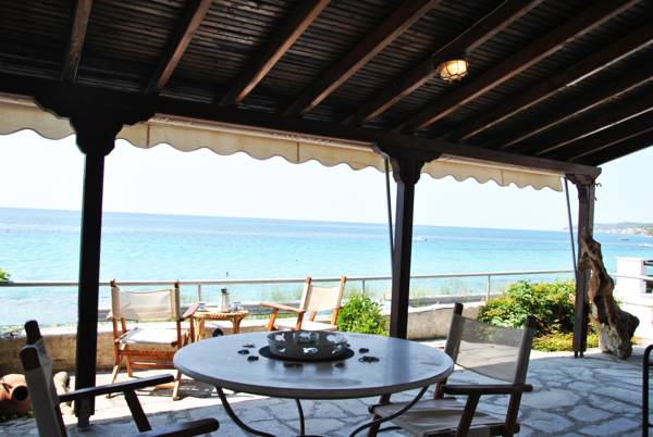 An outdoor terrace features a round dining table with decorative items, surrounded by wooden chairs. Beyond the terrace, clear blue water meets the shoreline, with gentle waves visible. The shaded area offers a relaxing vantage point overlooking the beach.
