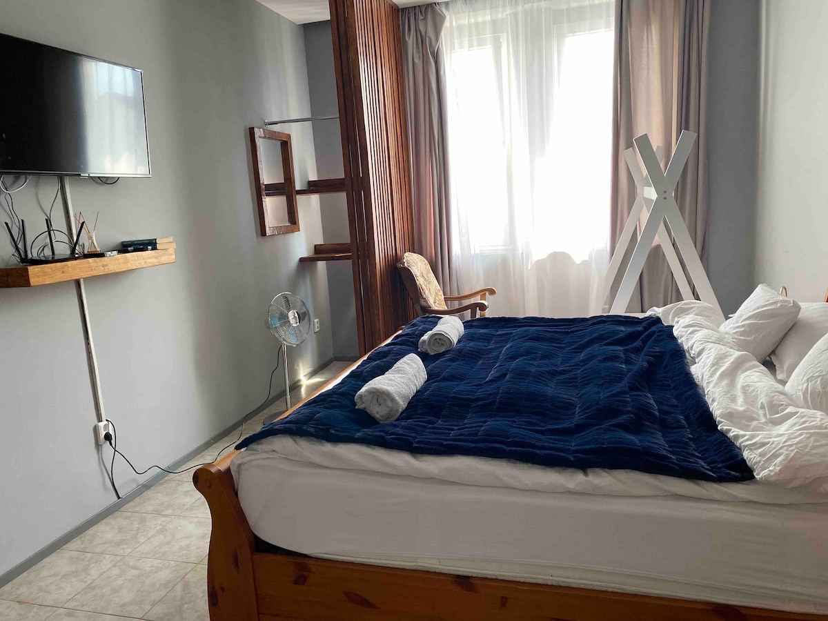 A sleeping area features a spacious bed layered with a blue bedspread and two rolled towels. A wooden chair and a fan are positioned nearby. Natural light enters through large windows, which are framed by soft curtains, complementing the neutral walls.