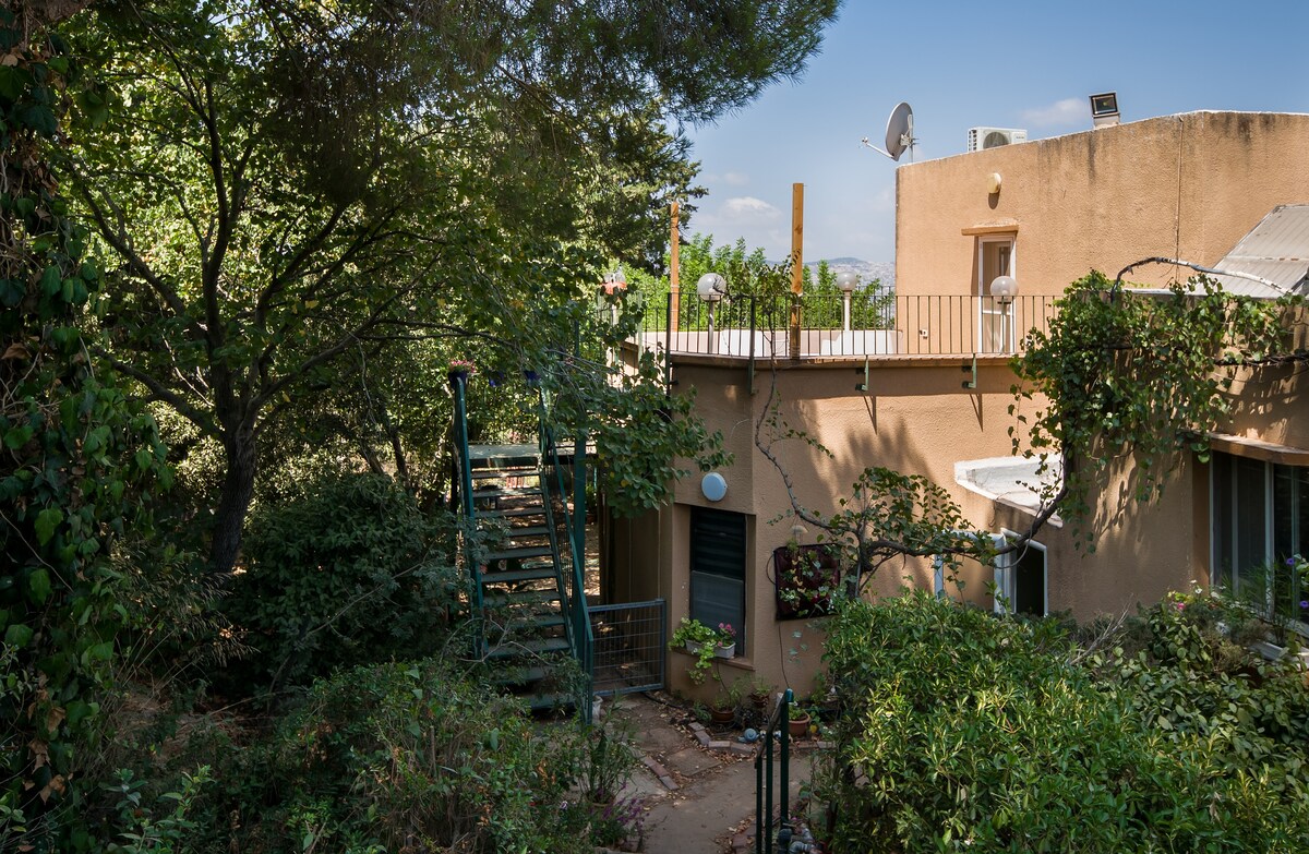 The view captures the exterior of a two-story building surrounded by lush greenery. A staircase leads to the upper level, with a terrace visible above. Soft sunlight filters through the trees, creating a serene atmosphere in the outdoor space.