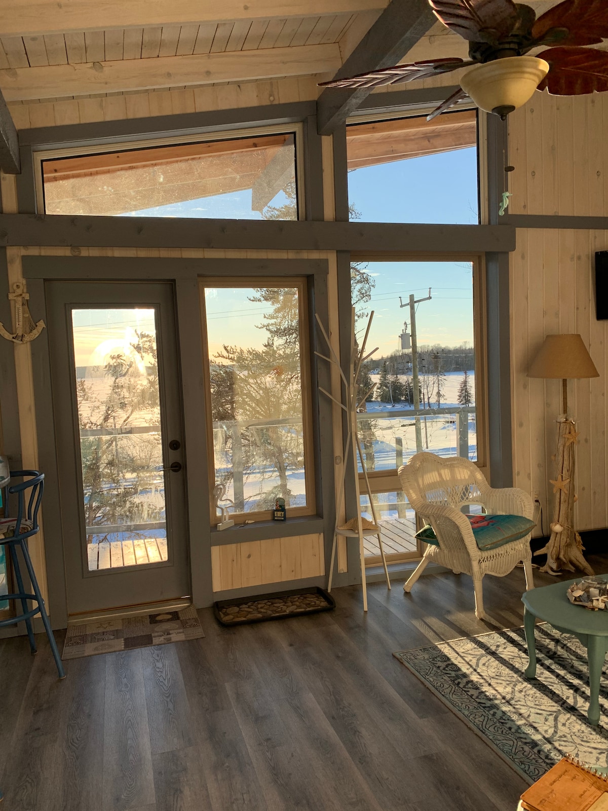 A bright and airy living space features large windows allowing natural light to flood in. A cozy seating area is positioned near the glass doors, offering views of the snowy landscape outside. The decor includes a tropical plant and a small table with decorative items.