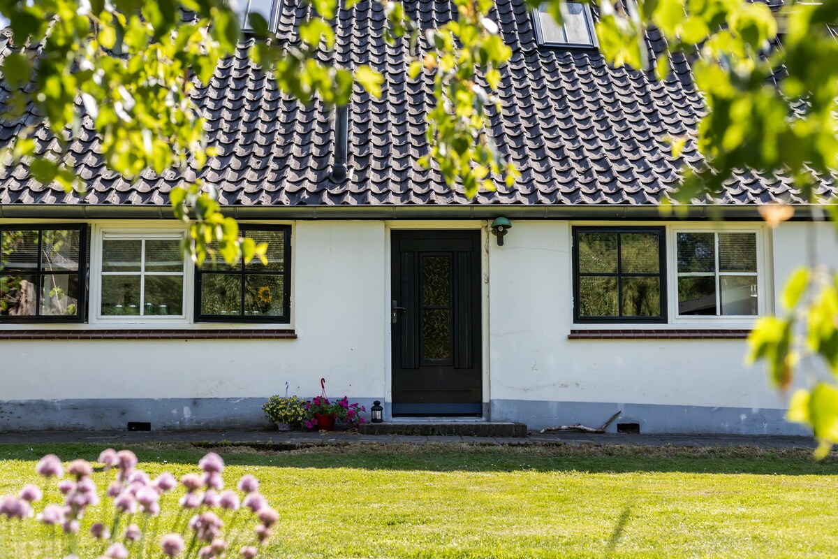 The exterior of the house features a tiled roof and a central entrance door framed by two large windows. A well-maintained lawn with flower beds is visible in the foreground, showcasing a welcoming approach to the property.