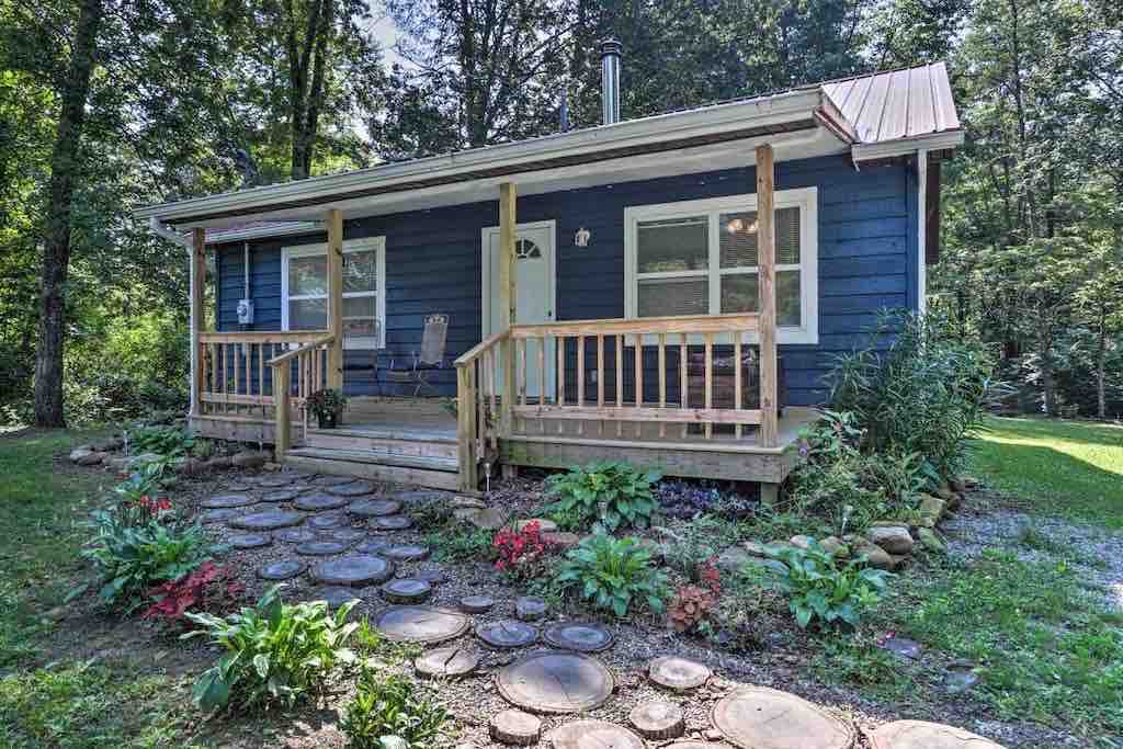 A charming cabin exterior is presented, featuring a welcoming porch with two chairs. A pathway of stepping stones leads through a lush green yard adorned with colorful flowers, while trees provide a natural backdrop.