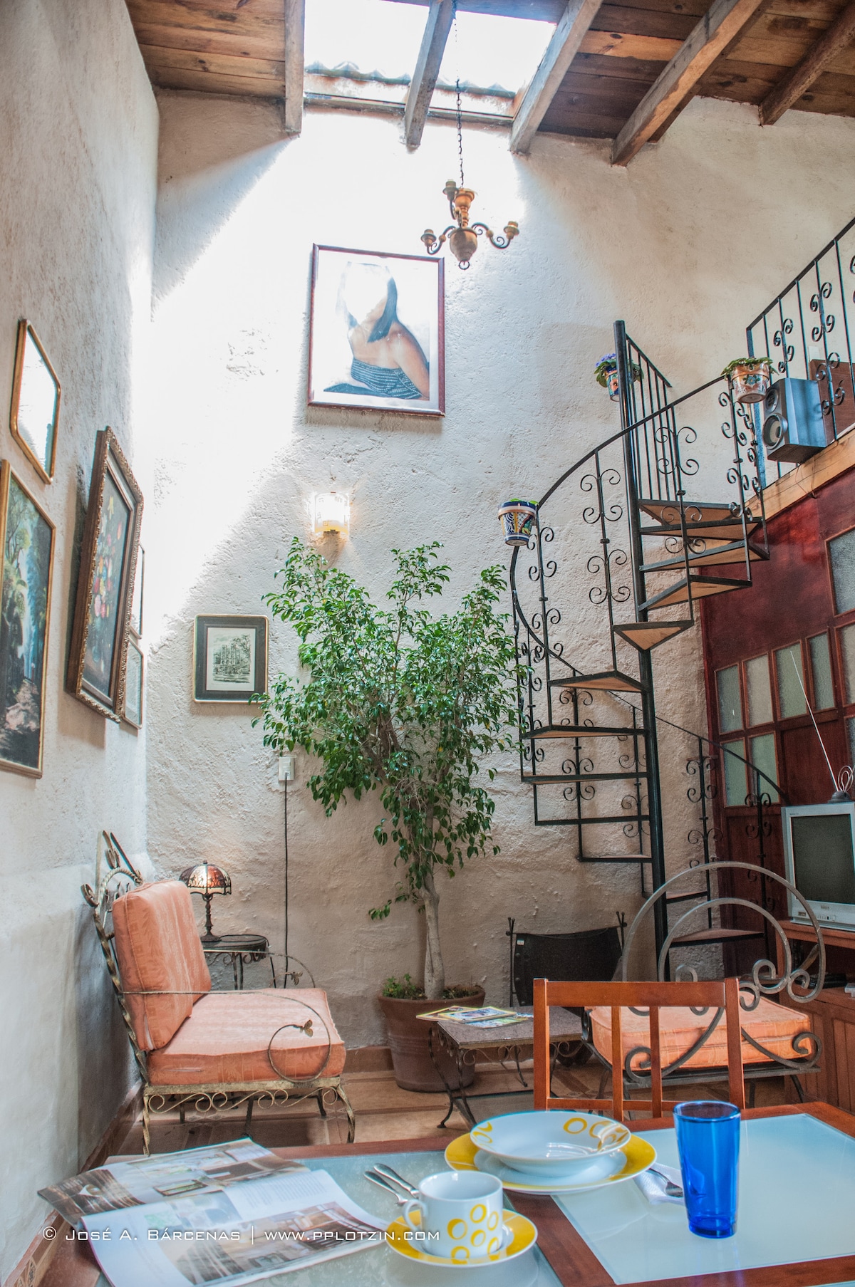 A welcoming living space features a spiral staircase leading to an upper level. Artwork adorns the textured walls, and a potted plant adds a touch of greenery. The dining area includes a wooden table set with dishes and glassware, surrounded by various seating arrangements.