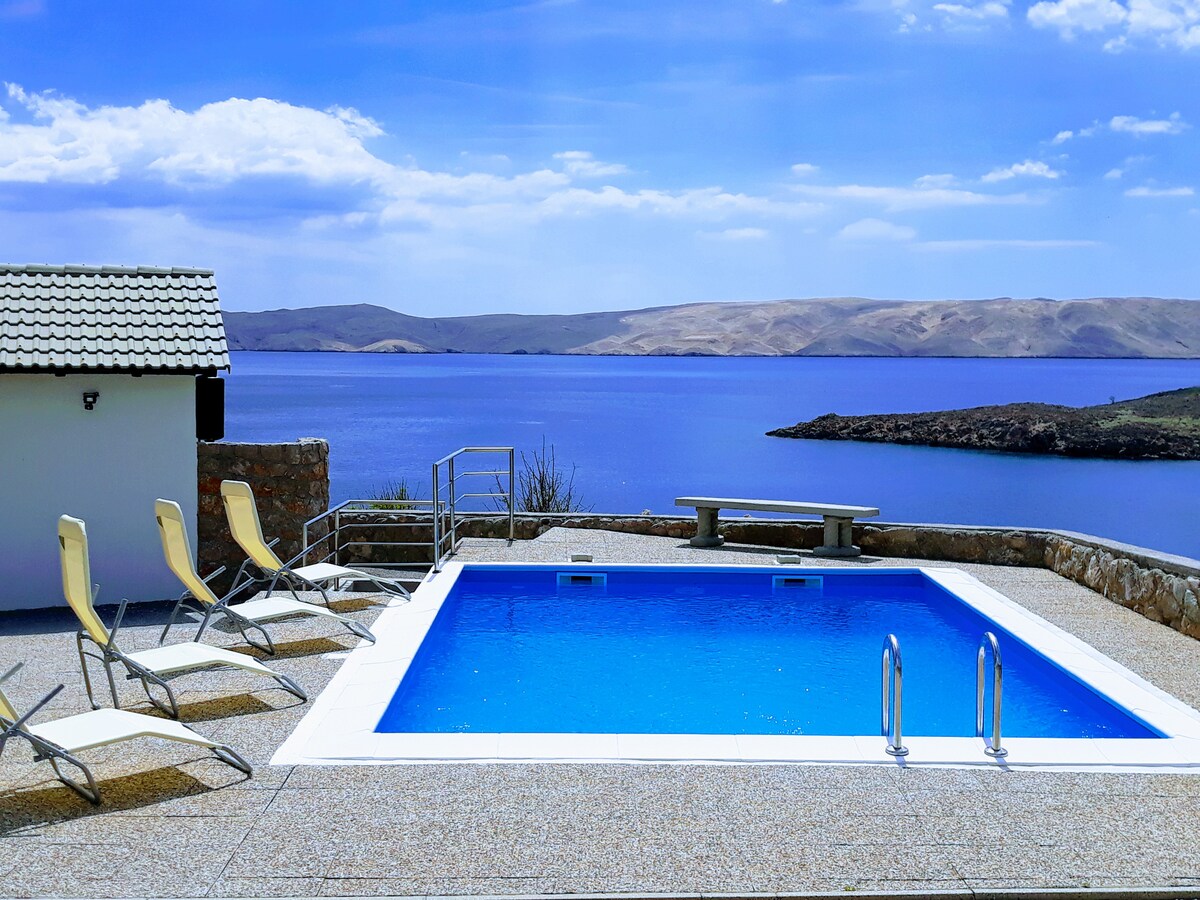 The outdoor area features a clear blue swimming pool surrounded by a sun terrace. Four lounge chairs are arranged along one side. A small building sits at the edge, while the view of the calm sea and distant hills enhances the serene environment.