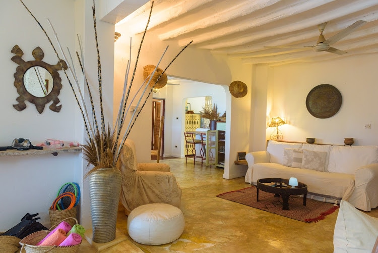 A welcoming living space features white furnishings and natural decor. A comfortable seating area is complemented by a low coffee table and decorative elements, such as a woven wall piece and potted plants. Light streams in from the entryway, enhancing the cozy atmosphere.
