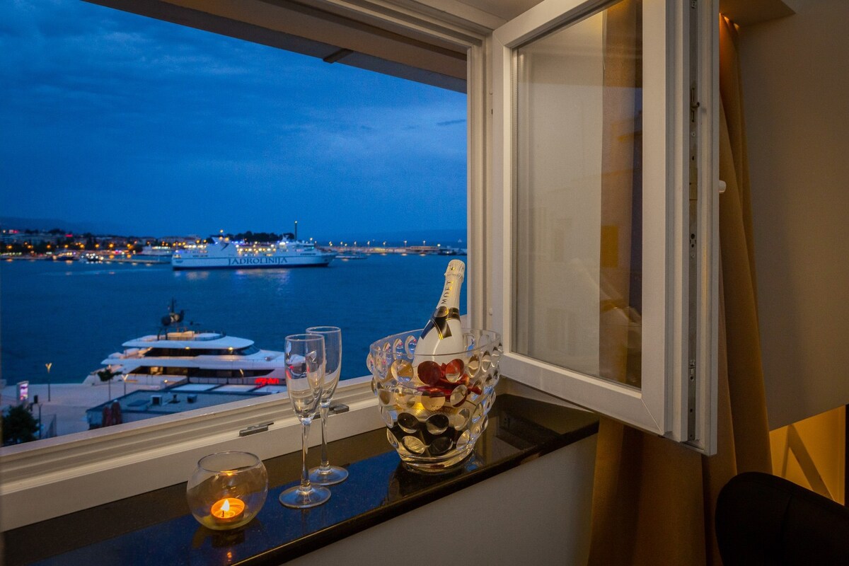 A window opens to a view of the harbor, showcasing boats gently bobbing in the water. A bottle of champagne and glasses sit on the windowsill, alongside a lit candle casting a warm glow. The early evening sky displays soft blue hues.