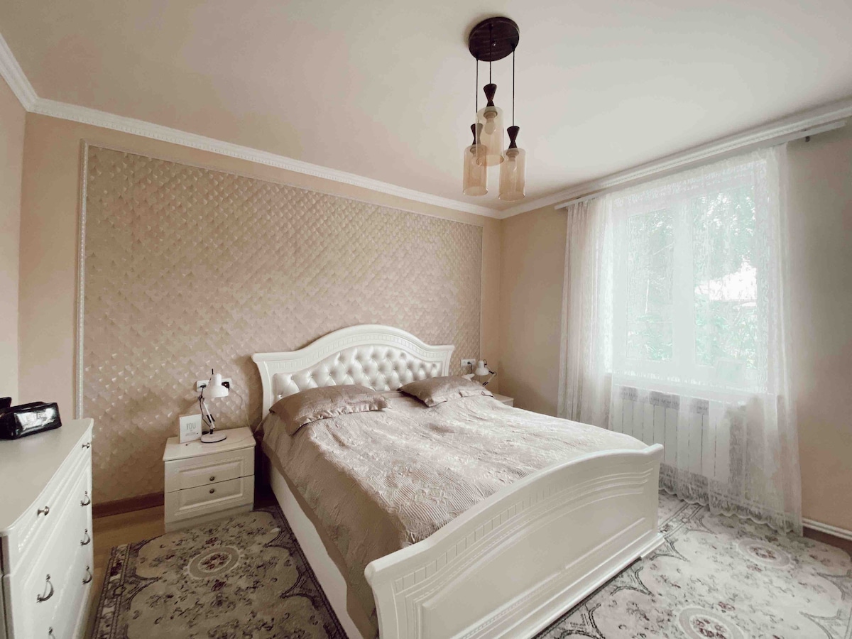A well-appointed bedroom features a spacious bed with elegant bedding, complemented by a decorative headboard. A window draped in sheer curtains allows natural light to fill the room, while a soft rug lies beneath. Two bedside tables provide additional space for personal items.